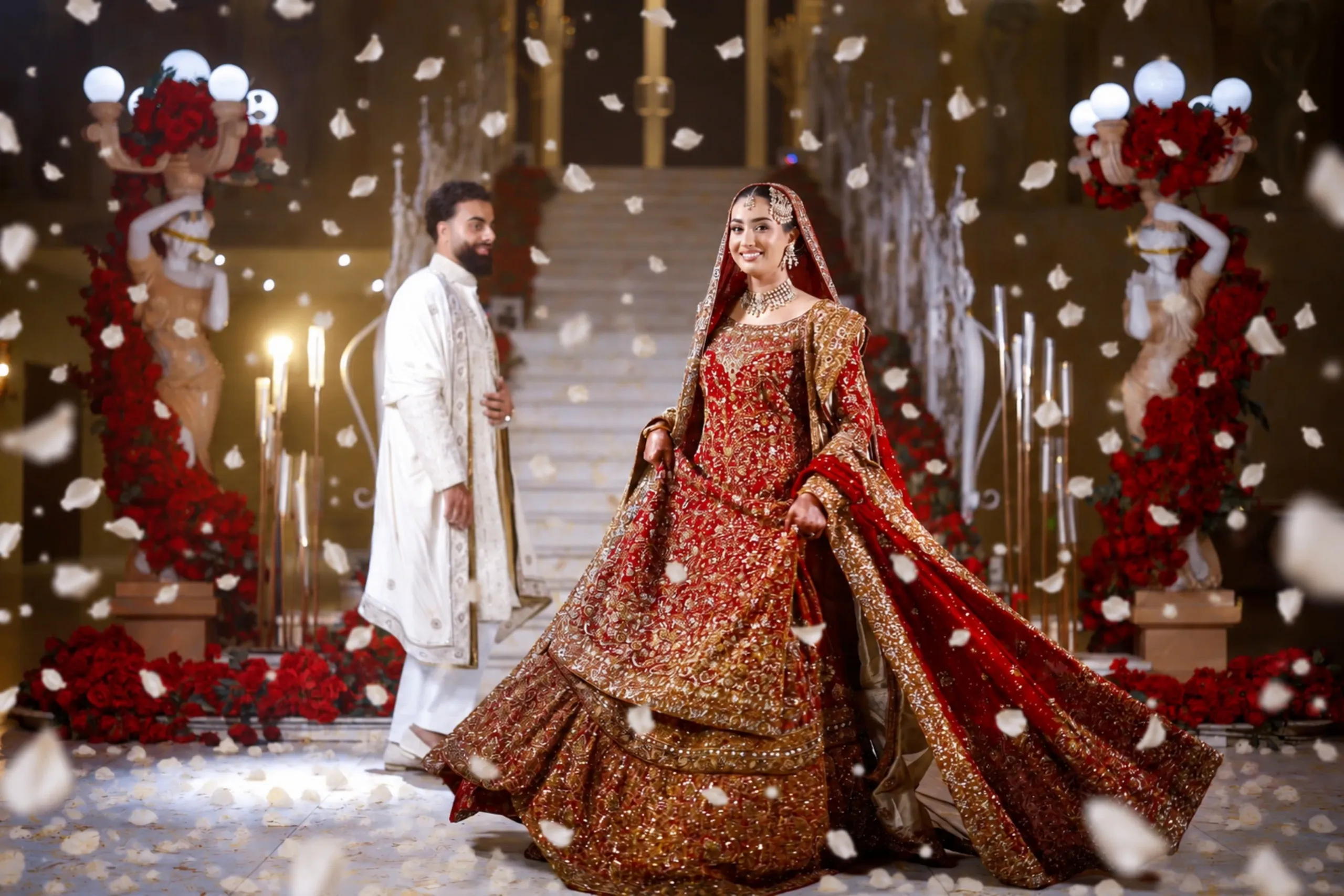 Bride twirling in red and gold on a rose-lined staircase as her groom looks on